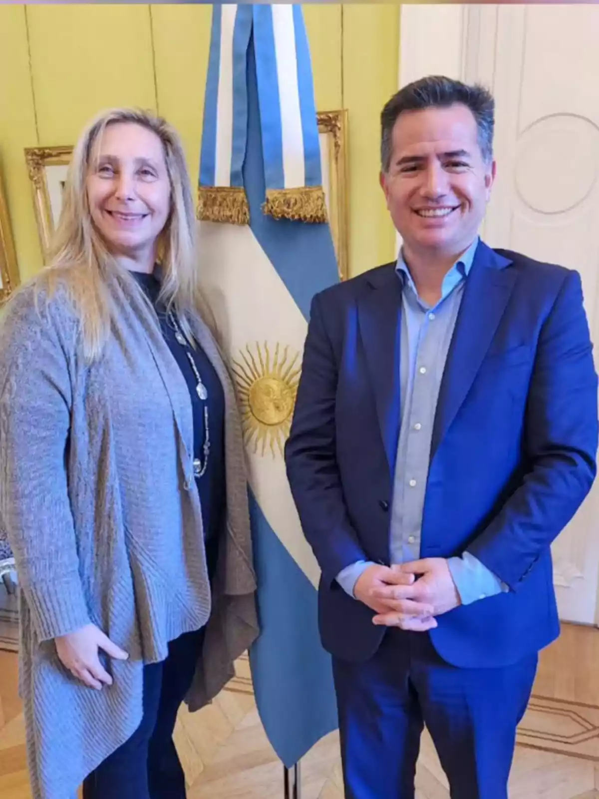 Dos personas sonrientes posan frente a una bandera de Argentina en un ambiente formal Dos personas sonrientes posan frente a una bandera de Argentina en un ambiente formal