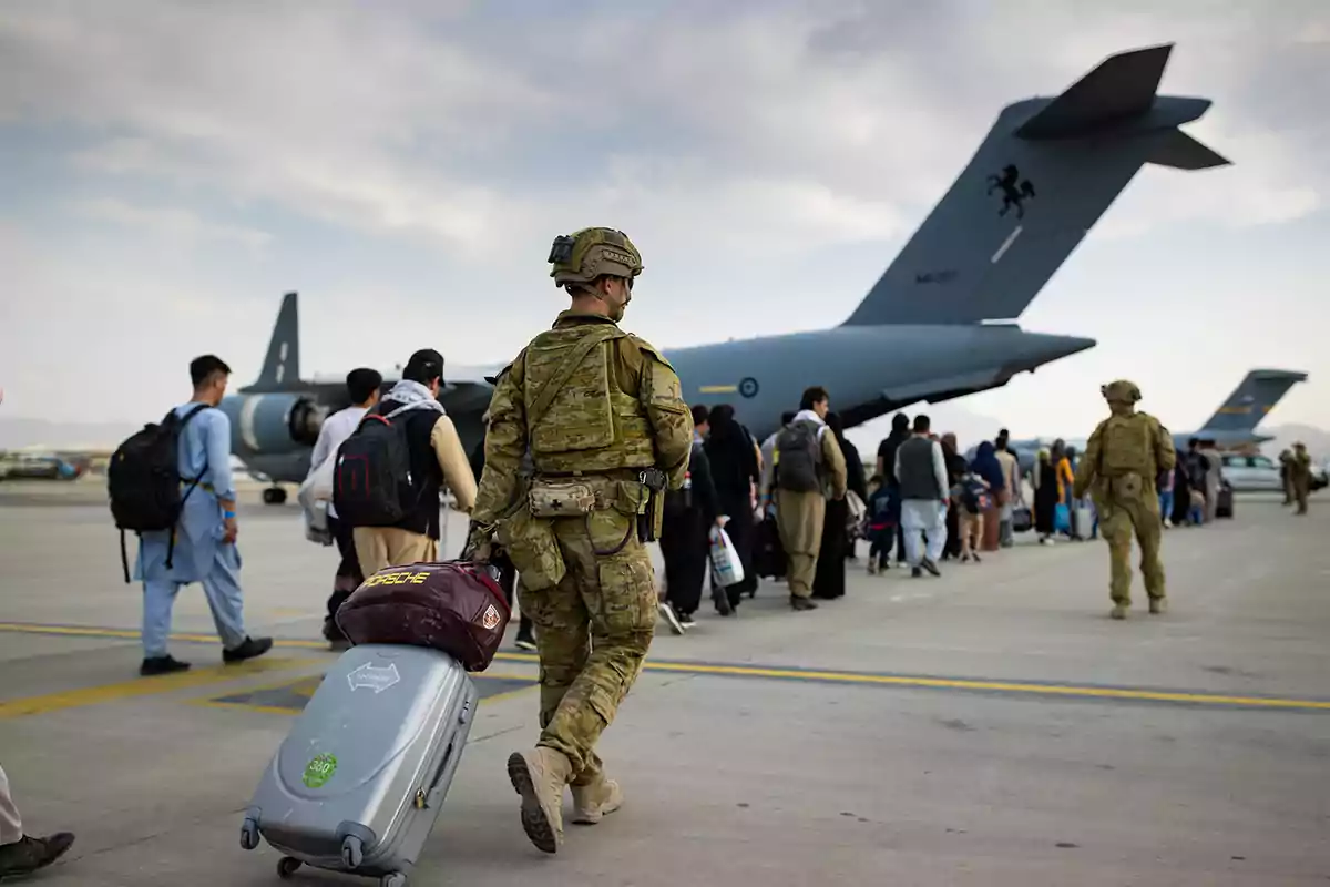 Soldados y civiles caminan hacia un avión militar en una pista de aterrizaje mientras uno de los soldados arrastra una maleta Soldados y civiles caminan hacia un avión militar en una pista de aterrizaje mientras uno de los soldados arrastra una maleta