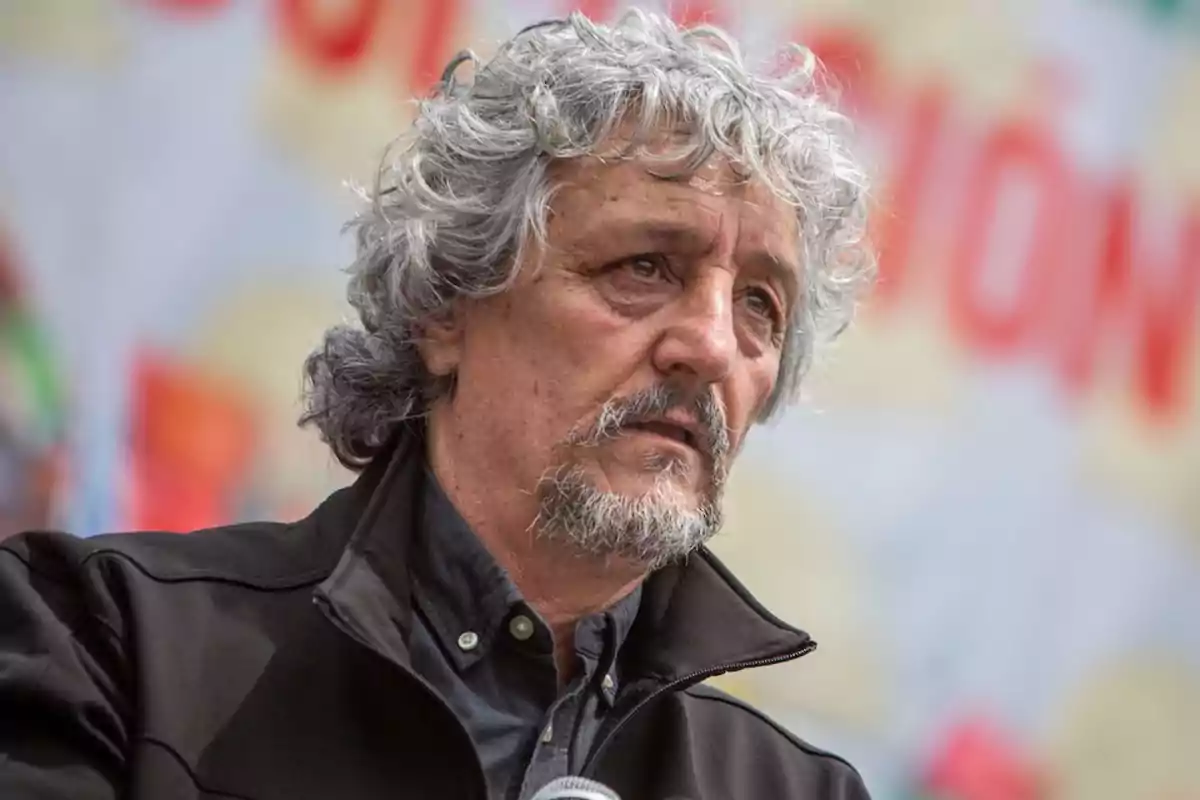 Hombre de cabello canoso y rizado con barba y chaqueta oscura mirando hacia el frente con expresión seria fondo desenfocado con colores claros y letras rojas Hombre de cabello canoso y rizado con barba y chaqueta oscura mirando hacia el frente con expresión seria fondo desenfocado con colores claros y letras rojas
