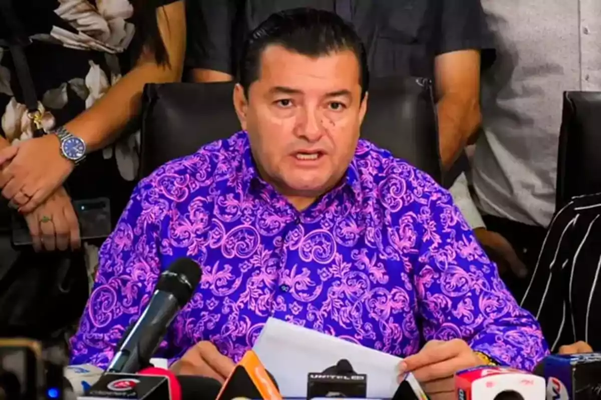 Hombre con camisa morada de estampado floral hablando frente a varios micrófonos en una conferencia de prensa rodeado de personas Hombre con camisa morada de estampado floral hablando frente a varios micrófonos en una conferencia de prensa rodeado de personas
