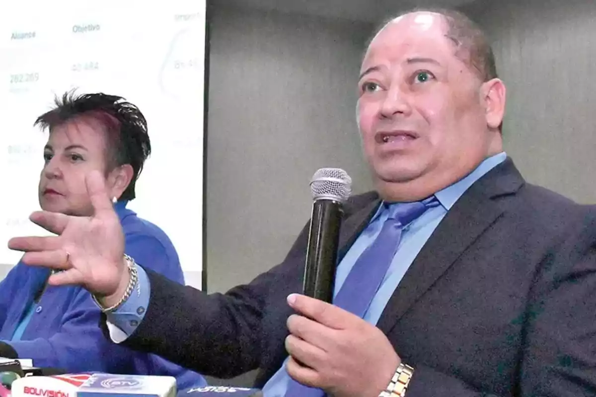 Un hombre con traje y corbata azul sostiene un micrófono mientras gesticula con la mano, a su lado una mujer también vestida de azul está sentada, ambos parecen estar en una conferencia o presentación.