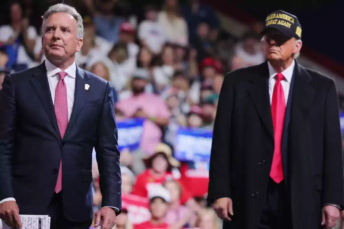 Two men in suits at a public event with a crowd of people in the background; one of them is wearing a black cap and a dark coat. Two men in suits at a public event with a crowd of people in the background; one of them is wearing a black cap and a dark coat.