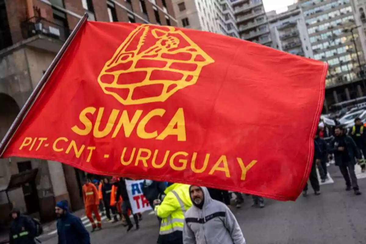 Red SUNCA union flag from Uruguay held during a demonstration on a street surrounded by buildings and people marching Red SUNCA union flag from Uruguay held during a demonstration on a street surrounded by buildings and people marching