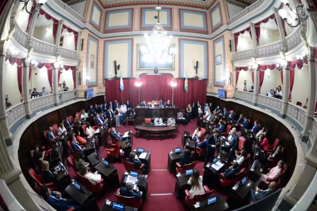 Panoramic view of a legislative chamber with numerous officials seated at their desks and an audience in the galleries, decorated with red curtains and a large chandelier in the center.