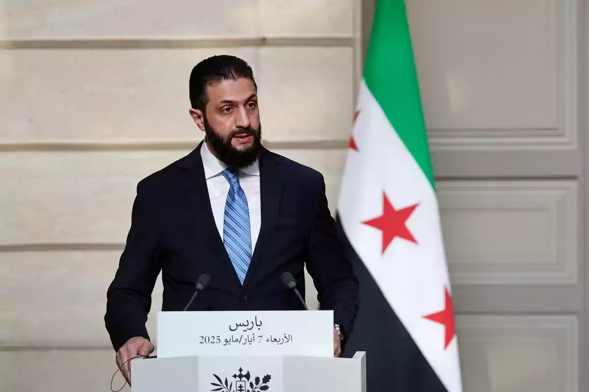 Hombre con barba y traje oscuro hablando en un podio con micrófonos y una bandera con estrellas rojas y fondo verde, blanco y negro detrás de él