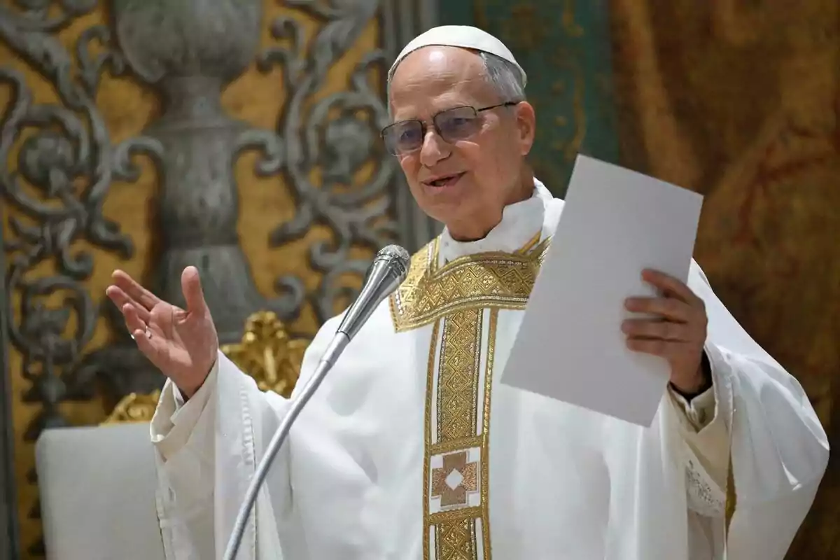 A priest dressed in white with gold details holds a paper and speaks into a microphone in an elegantly decorated setting. A priest dressed in white with gold details holds a paper and speaks into a microphone in an elegantly decorated setting.