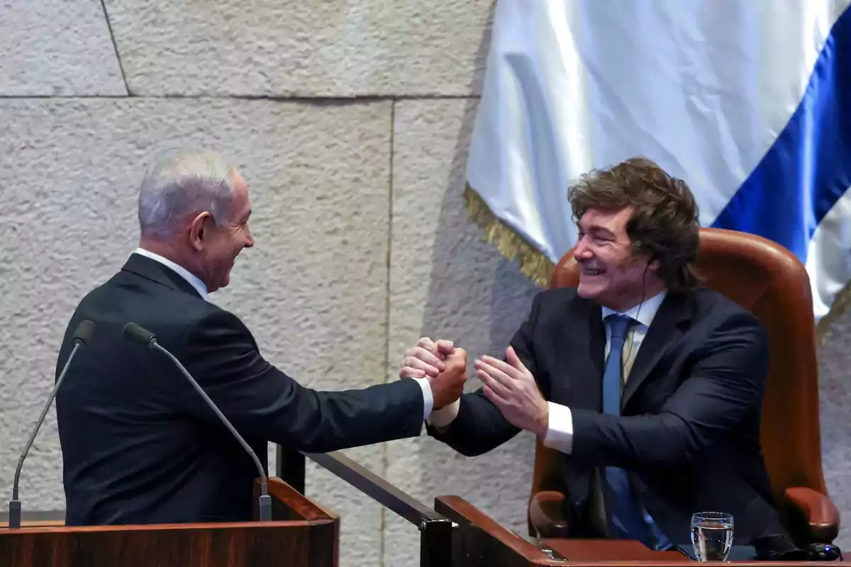 Two men in suits greet each other with a smile and a handshake in a formal setting next to a white and blue flag.