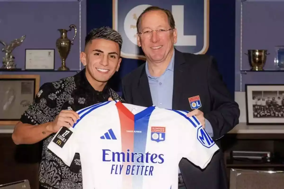 Dos personas sonríen mientras sostienen una camiseta del Olympique de Lyon en una sala con trofeos y fotografías al fondo Dos personas sonríen mientras sostienen una camiseta del Olympique de Lyon en una sala con trofeos y fotografías al fondo