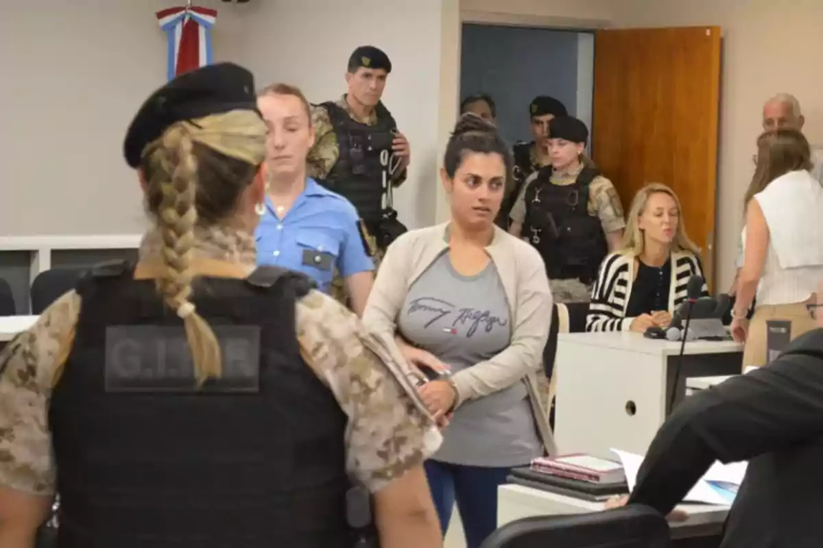 Una mujer esposada es escoltada por policías y personal de seguridad en una sala de audiencias mientras varias personas observan la escena Una mujer esposada es escoltada por policías y personal de seguridad en una sala de audiencias mientras varias personas observan la escena