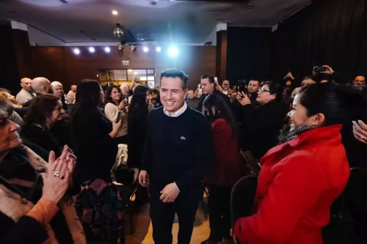 Un hombre sonriente camina entre un grupo de personas que lo aplauden y le toman fotos en un evento social en un salón iluminado. Un hombre sonriente camina entre un grupo de personas que lo aplauden y le toman fotos en un evento social en un salón iluminado.