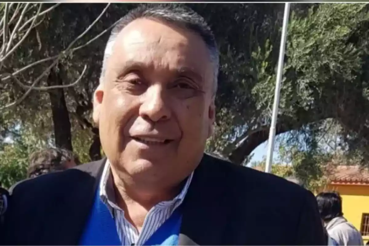 Hombre de cabello corto y canoso sonríe al aire libre con árboles y personas al fondo Hombre de cabello corto y canoso sonríe al aire libre con árboles y personas al fondo
