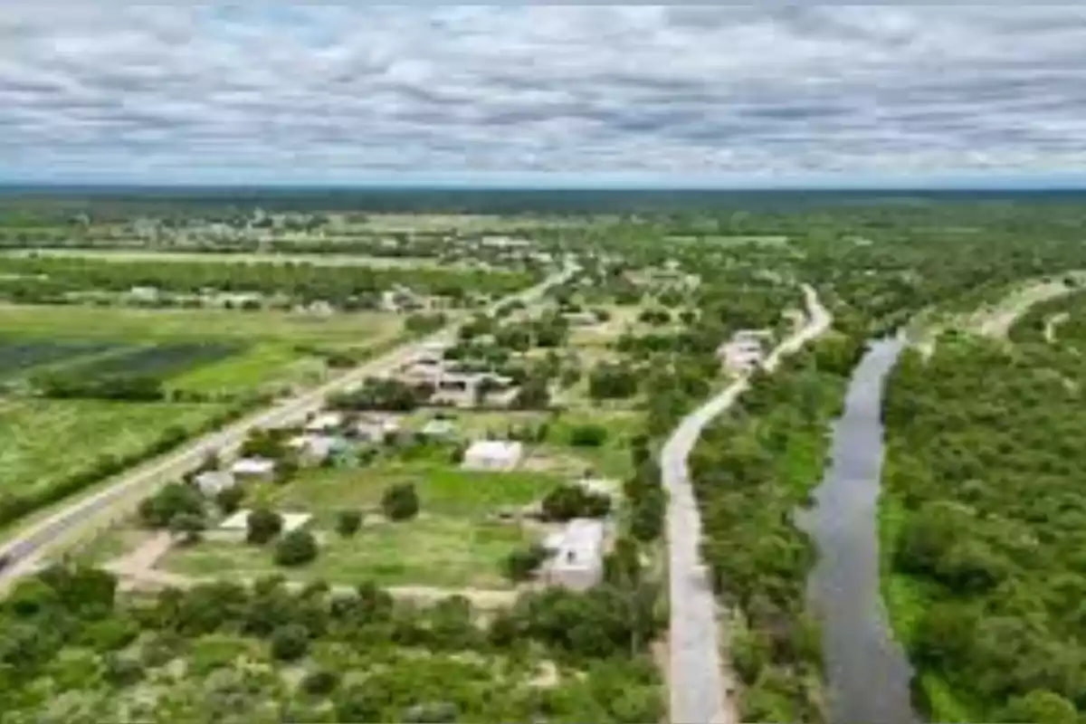 Vista aérea de un pequeño poblado rural rodeado de vegetación con un río y caminos que atraviesan la zona bajo un cielo nublado Vista aérea de un pequeño poblado rural rodeado de vegetación con un río y caminos que atraviesan la zona bajo un cielo nublado