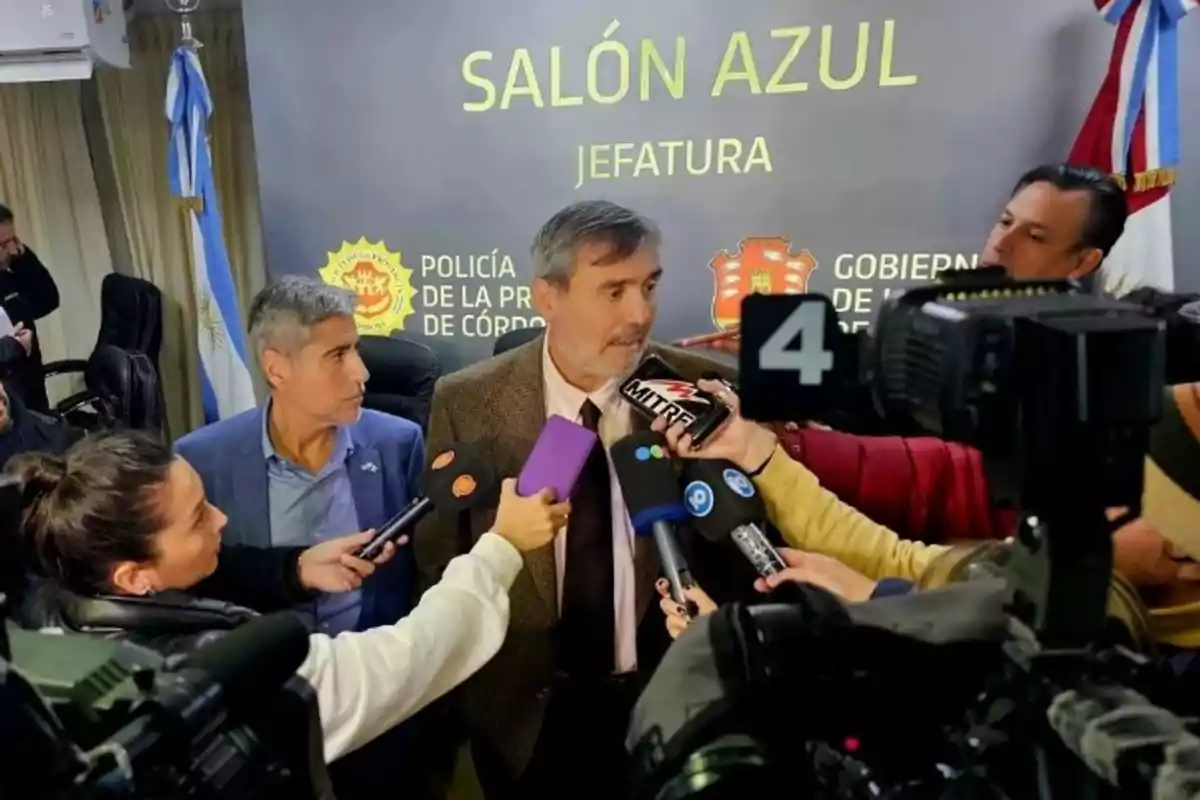 Un hombre de traje es entrevistado por varios periodistas en una sala con carteles de la policía de Córdoba y cámaras de televisión apuntando hacia él.