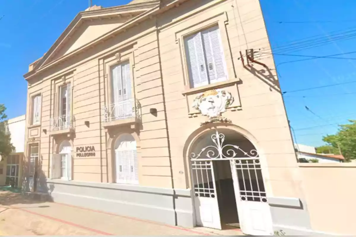 Edificio antiguo de color beige con ventanas y puertas blancas, un portón de hierro y un cartel que dice Policía Pellegrini bajo un cielo despejado. Edificio antiguo de color beige con ventanas y puertas blancas, un portón de hierro y un cartel que dice Policía Pellegrini bajo un cielo despejado.