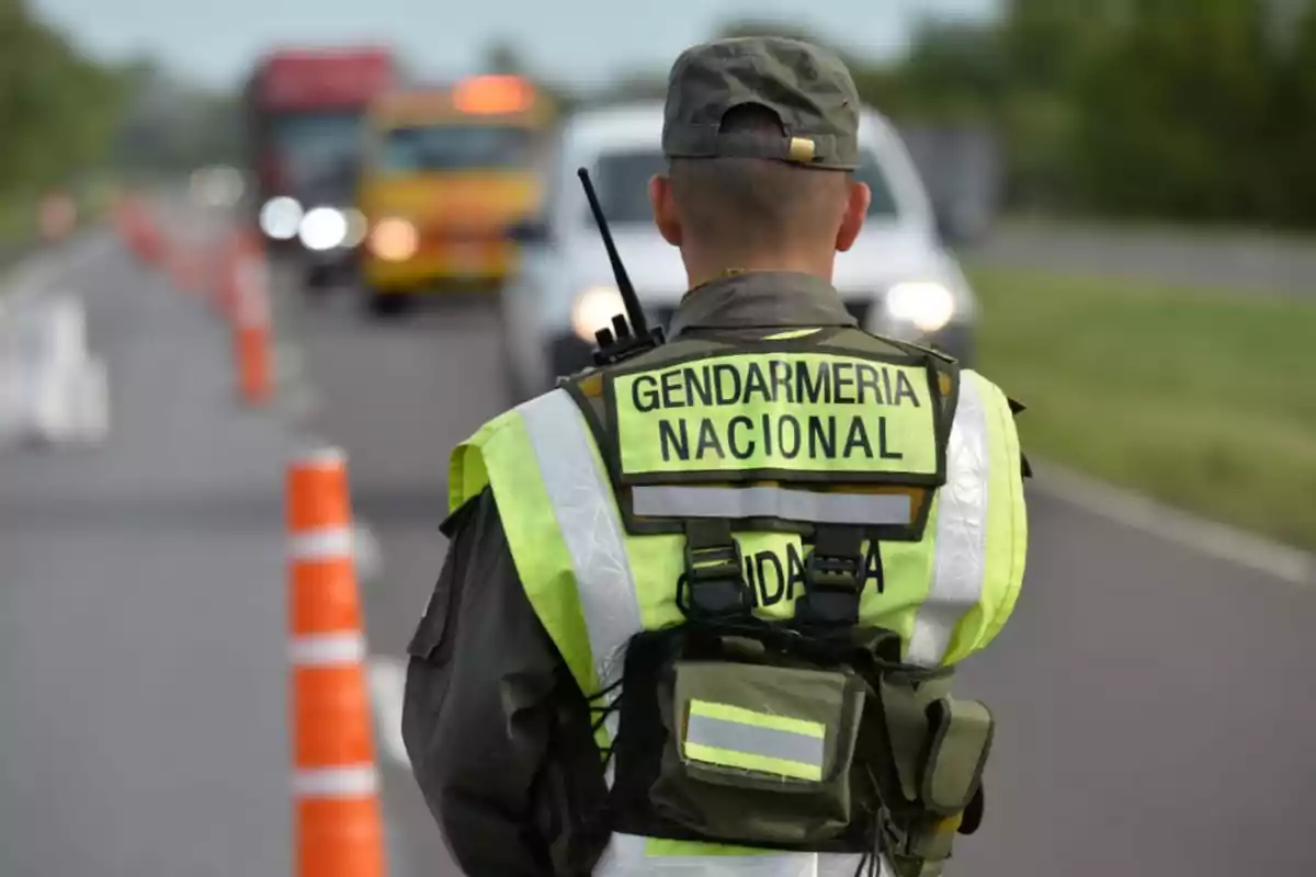 Agente de la Gendarmería Nacional de espaldas realizando un control vehicular en una carretera con conos naranjas y autos al fondo Agente de la Gendarmería Nacional de espaldas realizando un control vehicular en una carretera con conos naranjas y autos al fondo