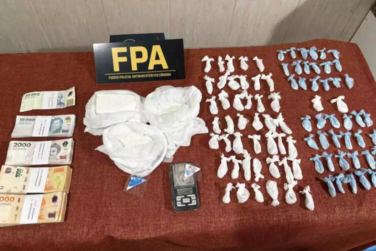 On a brown table, there are several bundles of bills, bags with white powder, a digital scale, small colored packages, and a sign from the Córdoba Anti-Narcotics Police Force. On a brown table, there are several bundles of bills, bags with white powder, a digital scale, small colored packages, and a sign from the Córdoba Anti-Narcotics Police Force.