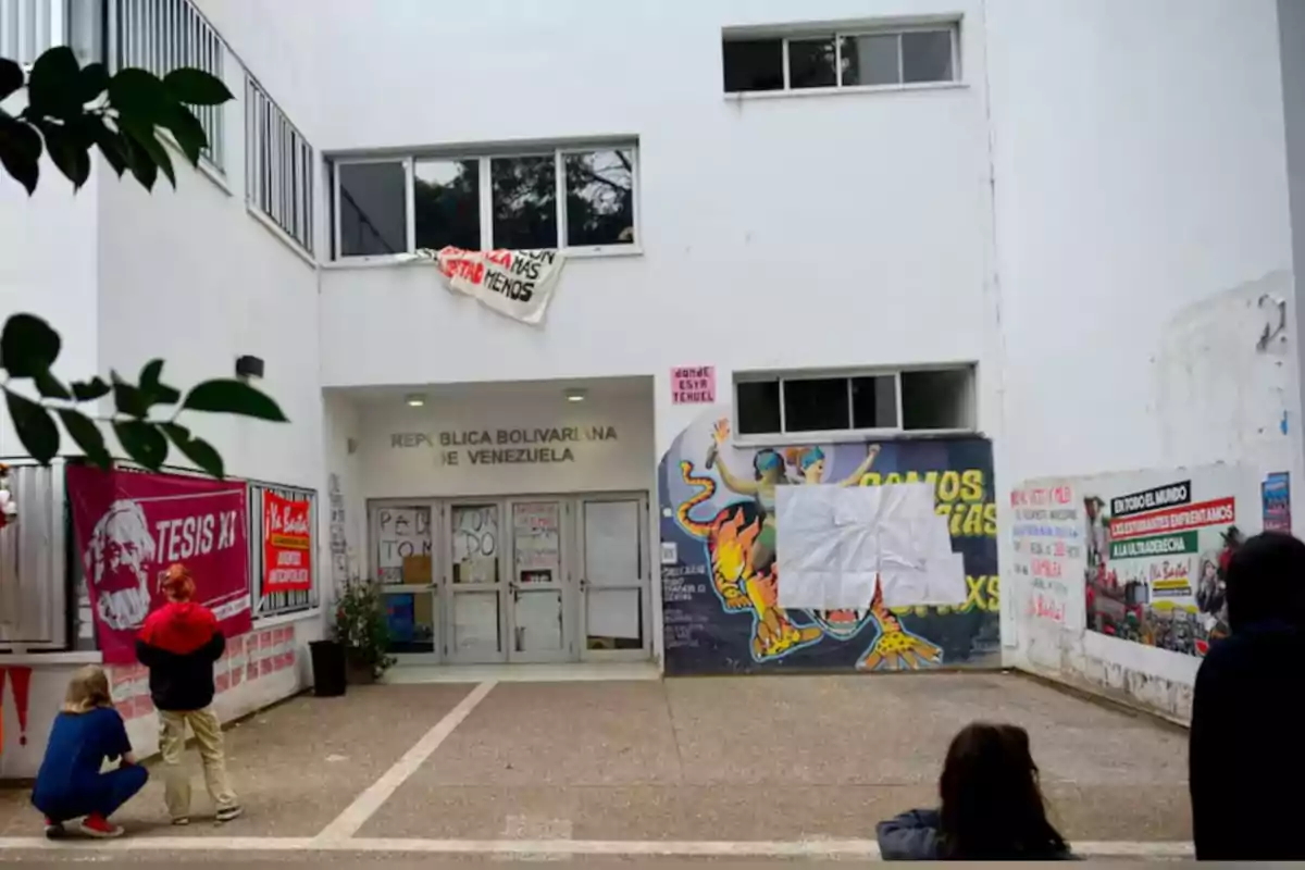 Facade of a white building with murals, posters, and banners, where several people are observing the place and the name Bolivarian Republic of Venezuela can be seen above the main entrance. Facade of a white building with murals, posters, and banners, where several people are observing the place and the name Bolivarian Republic of Venezuela can be seen above the main entrance.