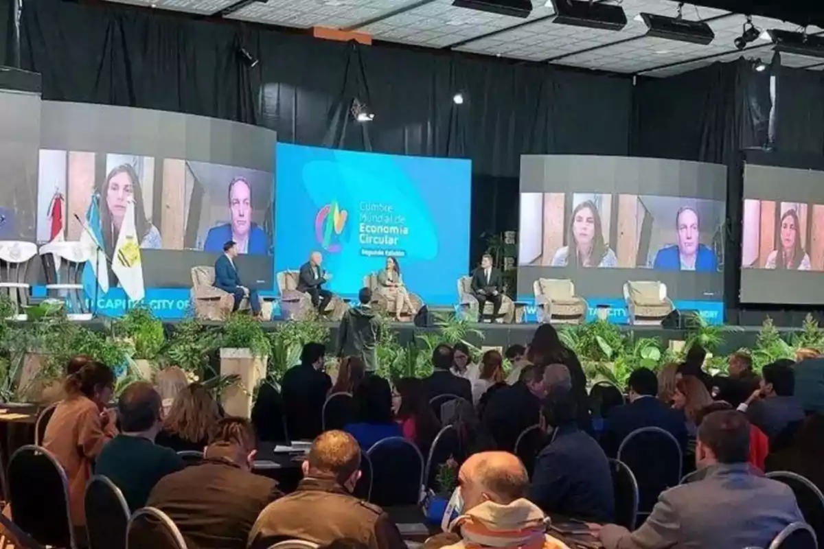 People attending a conference on circular economy with a panel of experts on stage and screens displaying video calls from remote participants People attending a conference on circular economy with a panel of experts on stage and screens displaying video calls from remote participants