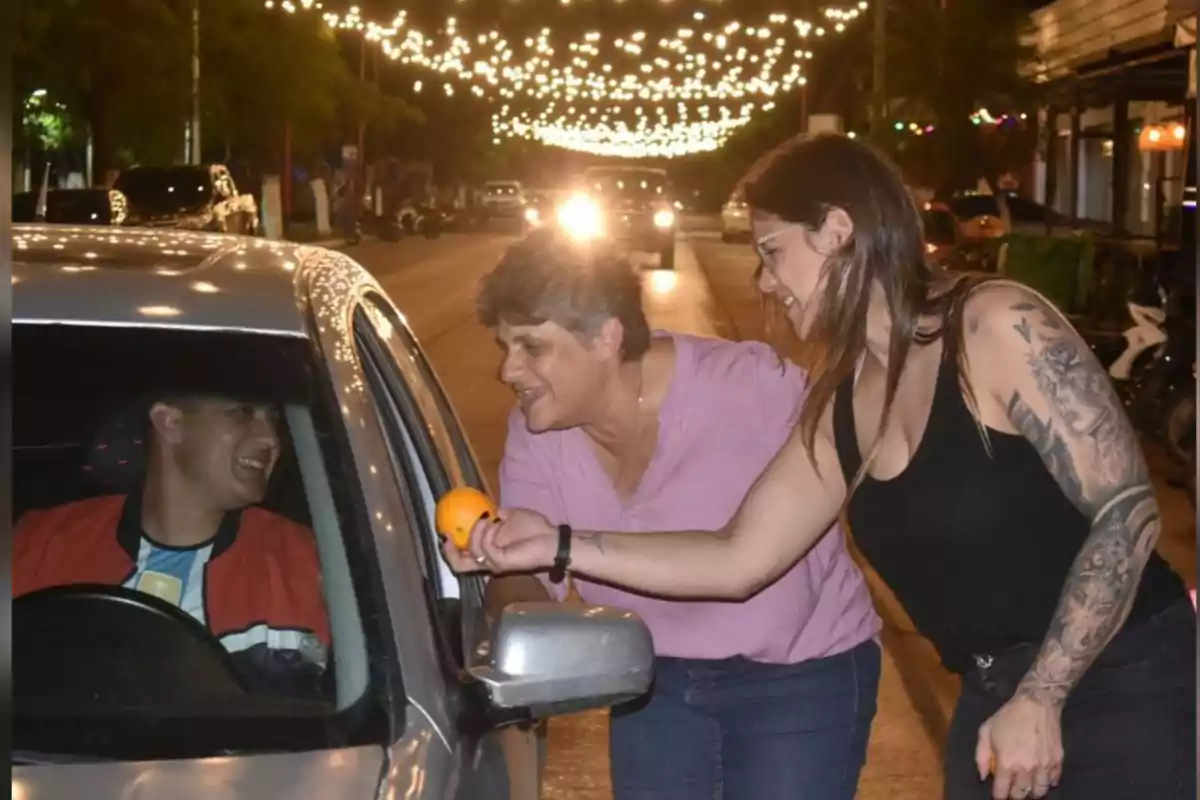 Dos mujeres sonrientes entregan una naranja a un hombre dentro de un auto en una calle iluminada por luces decorativas por la noche Dos mujeres sonrientes entregan una naranja a un hombre dentro de un auto en una calle iluminada por luces decorativas por la noche