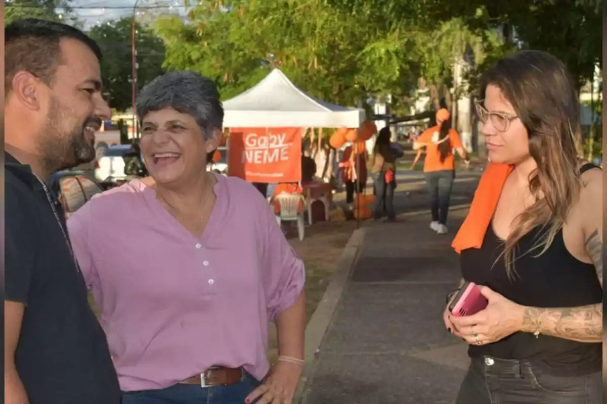Tres personas conversan y sonríen en un parque durante un evento al aire libre con un puesto de campaña política al fondo