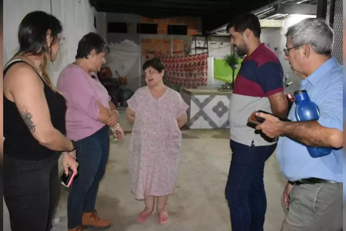 Cinco personas conversan en un espacio interior con paredes blancas y fondo de construcción, una de ellas está en el centro usando bata y sandalias, mientras las otras la rodean y escuchan atentamente. Cinco personas conversan en un espacio interior con paredes blancas y fondo de construcción, una de ellas está en el centro usando bata y sandalias, mientras las otras la rodean y escuchan atentamente.