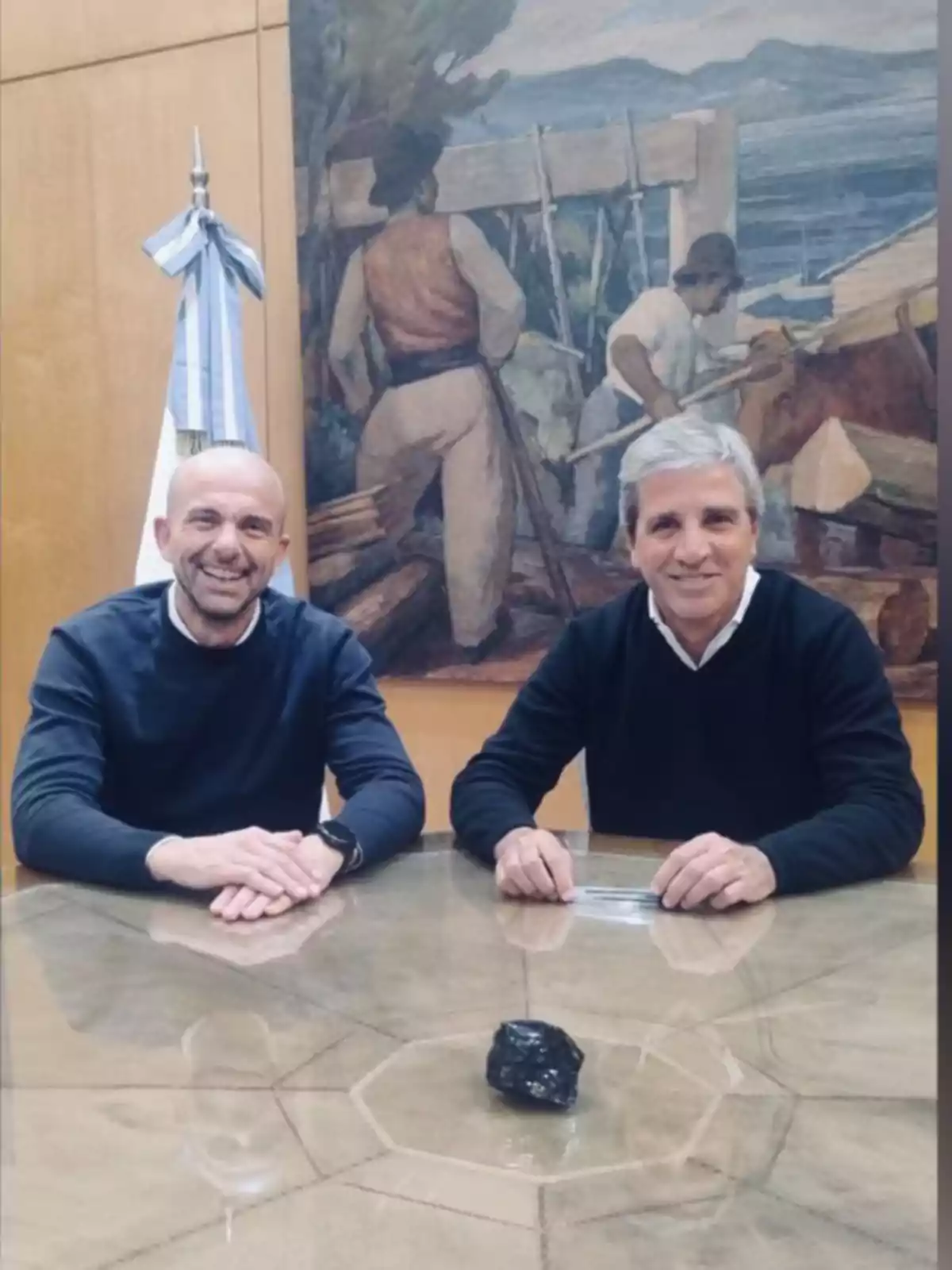 Two men sitting at a glass table with a black stone in the center and an Argentine flag in the background next to a mural of workers. Two men sitting at a glass table with a black stone in the center and an Argentine flag in the background next to a mural of workers.