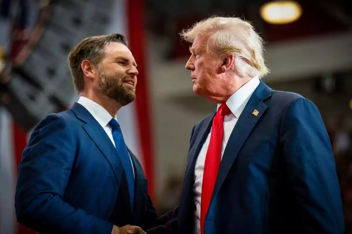Two men in suits shake hands and look at each other during an event in an indoor venue.