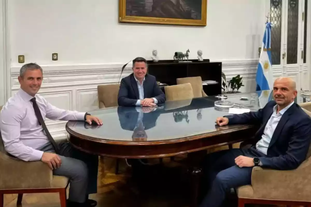 Three men in suits sitting around a glass table in a stylish office with an Argentine flag in the background Three men in suits sitting around a glass table in a stylish office with an Argentine flag in the background
