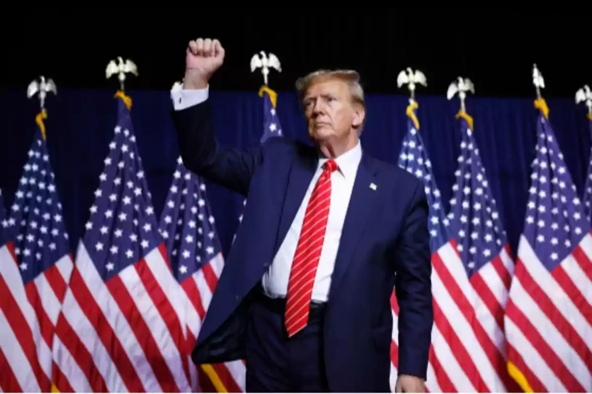 Man in a blue suit with a red tie raising his fist in front of several American flags Man in a blue suit with a red tie raising his fist in front of several American flags