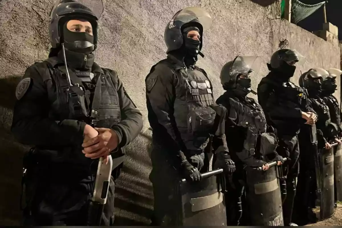 Grupo de policías antidisturbios con equipo de protección y escudos formados junto a una pared