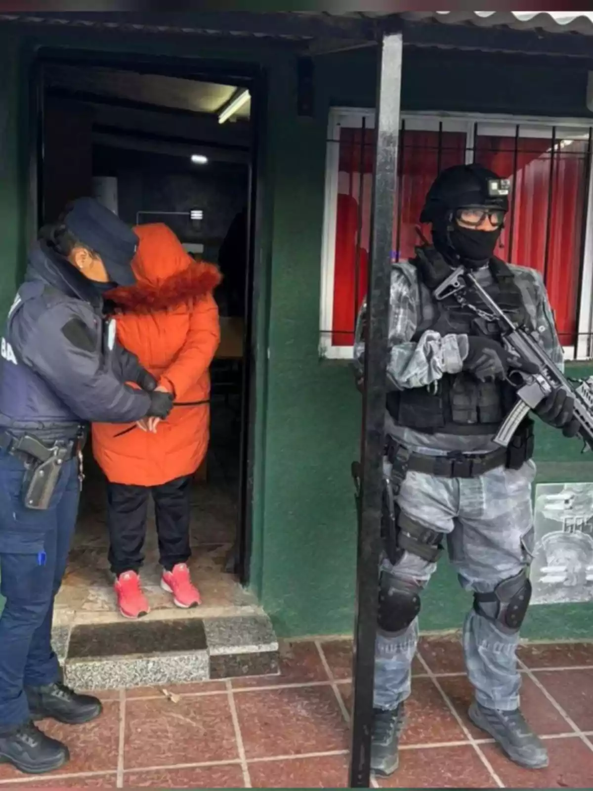 Una persona con abrigo naranja es escoltada por una policía mientras un agente fuertemente armado vigila la entrada de una casa