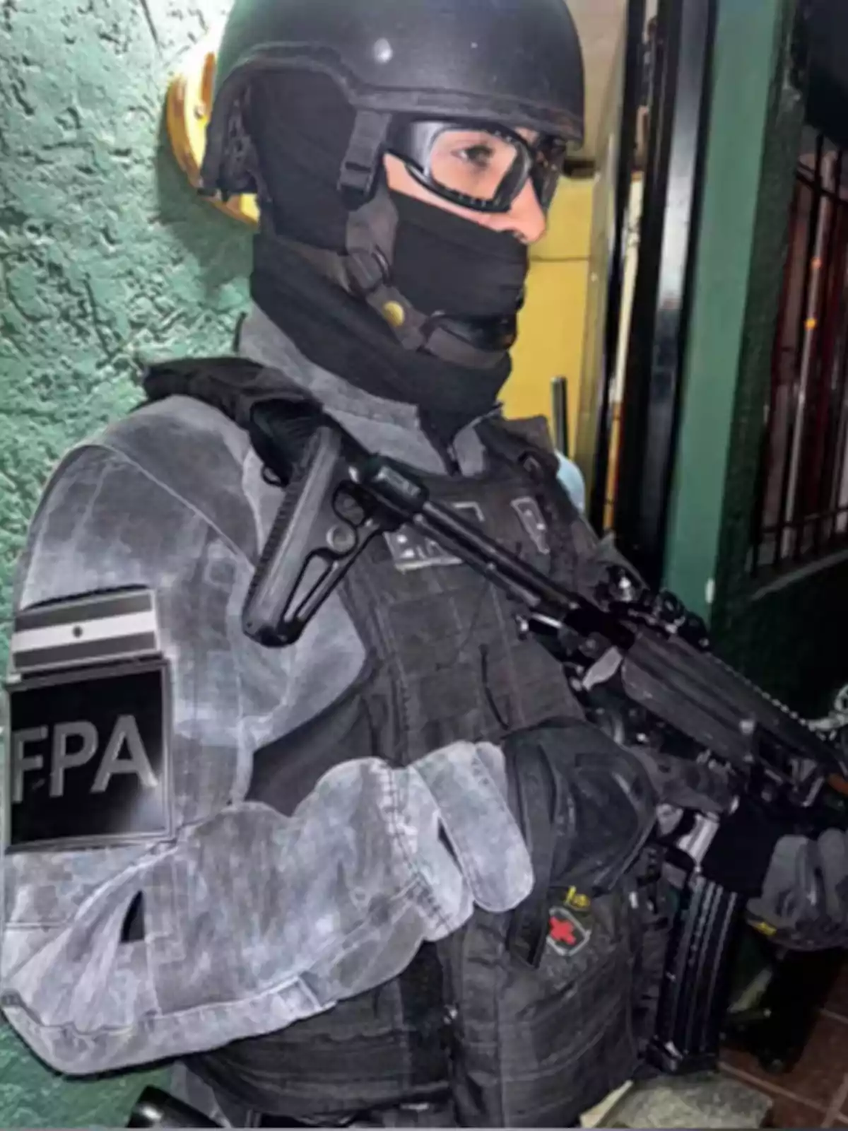Agente de fuerzas especiales con uniforme táctico y casco sosteniendo un arma frente a una pared verde