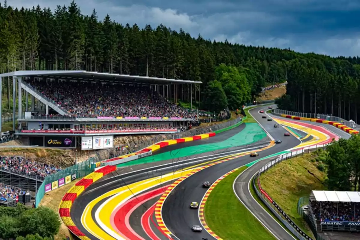 Panoramic view of a racetrack surrounded by trees, with grandstands full of spectators and several cars competing on the track. Panoramic view of a racetrack surrounded by trees, with grandstands full of spectators and several cars competing on the track.