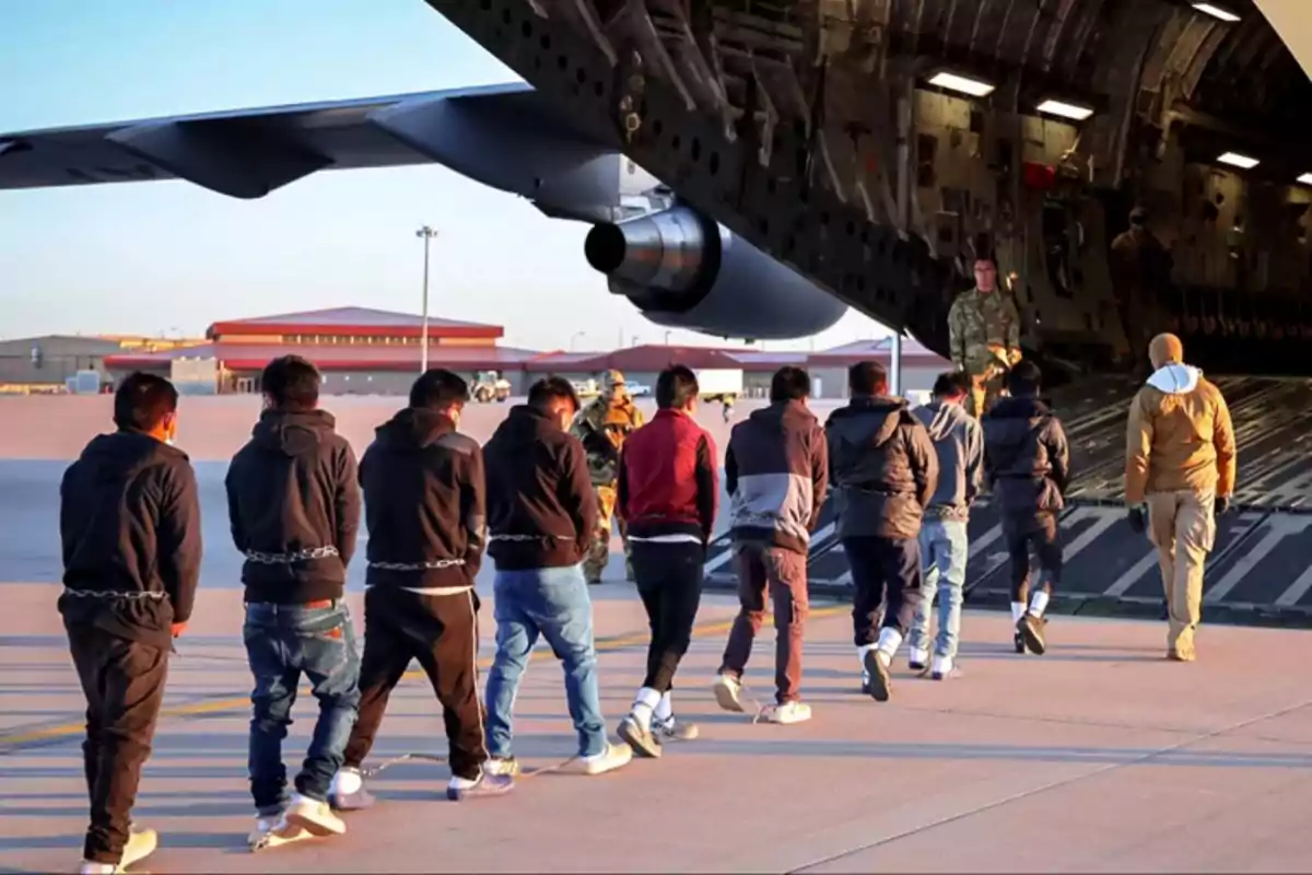 Un grupo de personas esposadas camina en fila hacia la rampa de un avión militar escoltados por personal uniformado en una pista de aeropuerto. Un grupo de personas esposadas camina en fila hacia la rampa de un avión militar escoltados por personal uniformado en una pista de aeropuerto.