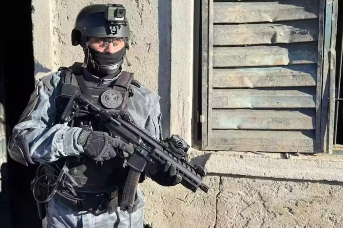 Un agente táctico con casco y equipo de protección sostiene un arma frente a una pared de concreto junto a una ventana vieja Un agente táctico con casco y equipo de protección sostiene un arma frente a una pared de concreto junto a una ventana vieja