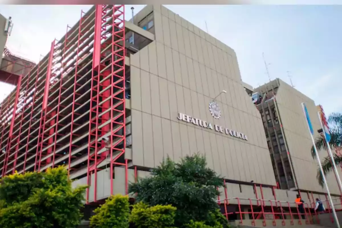 Edificio grande de la Jefatura de Policía con estructura moderna y detalles en color rojo rodeado de árboles y banderas Edificio grande de la Jefatura de Policía con estructura moderna y detalles en color rojo rodeado de árboles y banderas