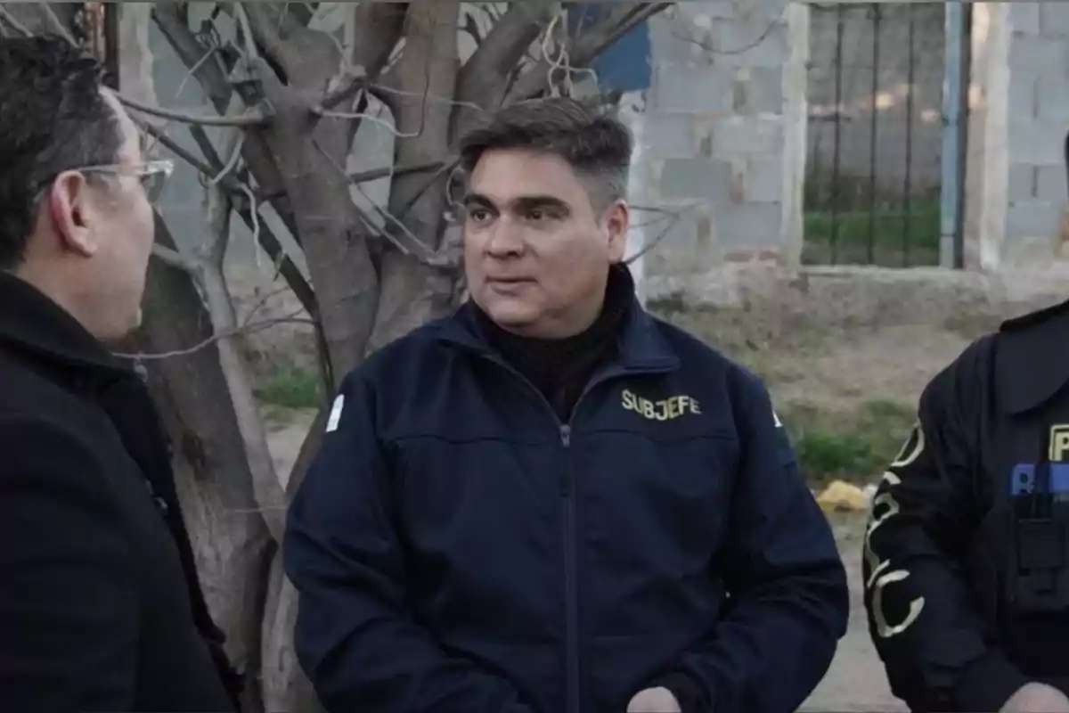 Tres hombres conversan al aire libre, uno de ellos lleva una chaqueta con la palabra subjefe y otro viste uniforme policial Tres hombres conversan al aire libre, uno de ellos lleva una chaqueta con la palabra subjefe y otro viste uniforme policial
