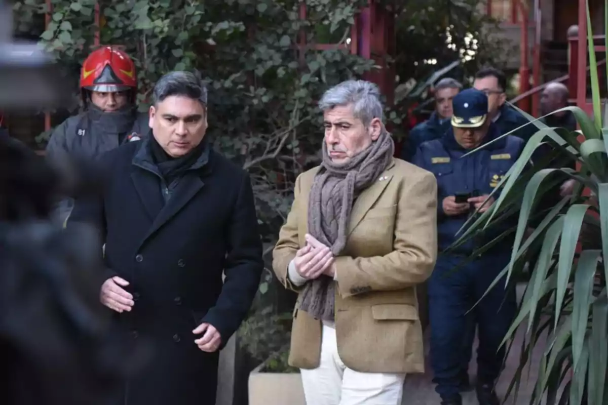 Dos hombres caminan juntos en primer plano, uno con abrigo negro y el otro con saco marrón y bufanda, mientras al fondo se observan bomberos y personal de seguridad Dos hombres caminan juntos en primer plano, uno con abrigo negro y el otro con saco marrón y bufanda, mientras al fondo se observan bomberos y personal de seguridad