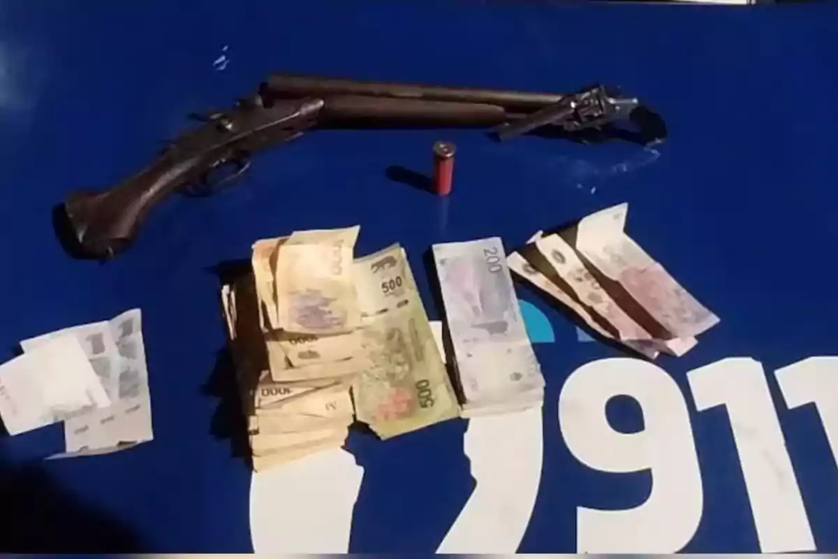 Escopeta antigua, cartucho y varios fajos de billetes argentinos sobre una superficie azul con el número 911 en blanco. Escopeta antigua, cartucho y varios fajos de billetes argentinos sobre una superficie azul con el número 911 en blanco.