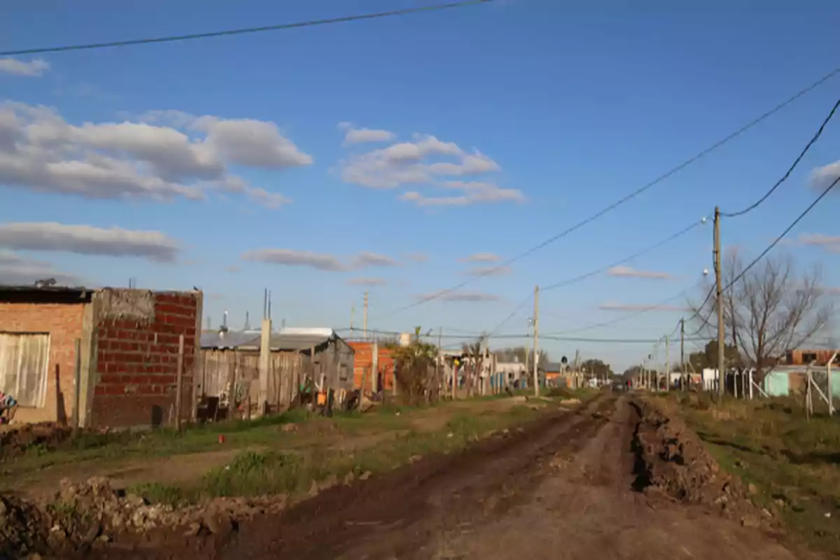 Calle de tierra en un barrio con casas humildes y postes de luz bajo un cielo azul con nubes dispersas