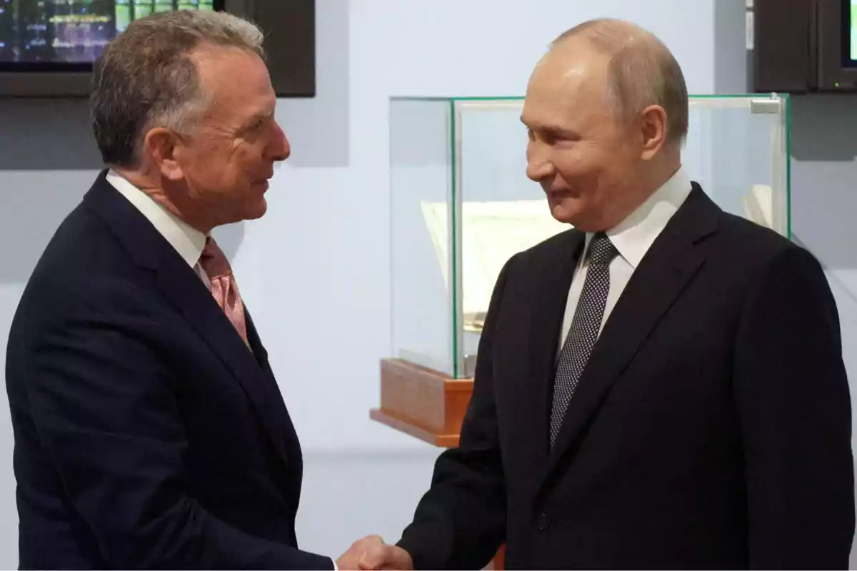 Two men in suits shake hands and smile at each other in a formal setting. Two men in suits shake hands and smile at each other in a formal setting.