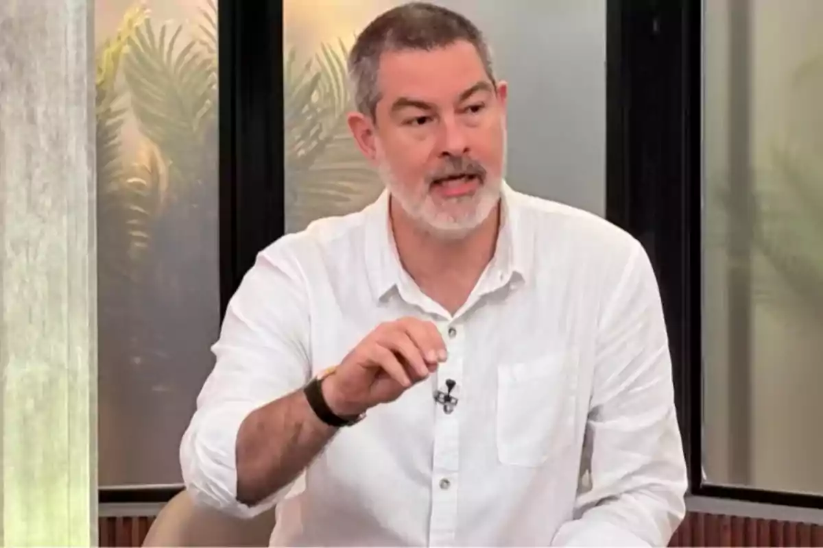 Short-haired, gray-bearded man wearing a white jersey while speaking and gesturing in an indoor setting with plants in the background Short-haired, gray-bearded man wearing a white jersey while speaking and gesturing in an indoor setting with plants in the background