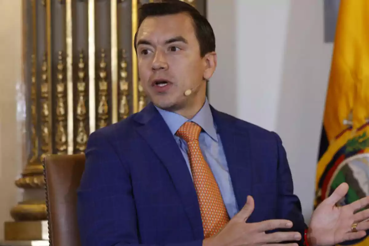 Hombre con traje azul y corbata naranja hablando con un micrófono de diadema en un entorno formal con una bandera de Ecuador al fondo