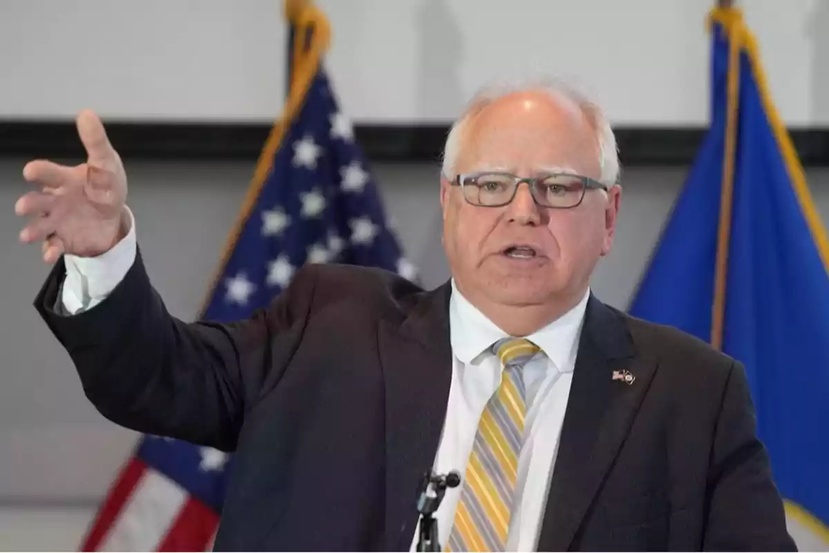 Gray-haired man with glasses speaking in public with flags in the background, wearing a dark suit and a striped tie.