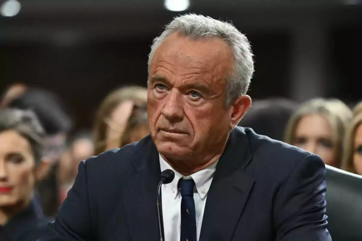 Gray-haired man with a serious expression wearing a dark suit and tie, seated in front of a microphone in a formal setting with blurred people in the background Gray-haired man with a serious expression wearing a dark suit and tie, seated in front of a microphone in a formal setting with blurred people in the background