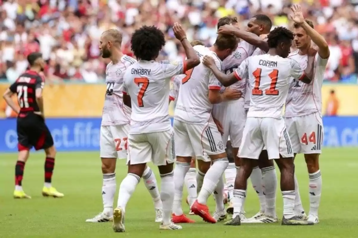 Players from a soccer team in white uniforms celebrate a goal on the field while an opponent in a black and red uniform watches from a distance. Players from a soccer team in white uniforms celebrate a goal on the field while an opponent in a black and red uniform watches from a distance.