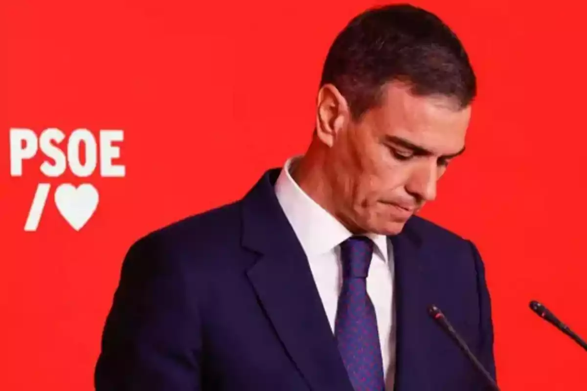 Hombre con traje oscuro y corbata mirando hacia abajo frente a un fondo rojo con el logo de PSOE y un corazón blanco Hombre con traje oscuro y corbata mirando hacia abajo frente a un fondo rojo con el logo de PSOE y un corazón blanco