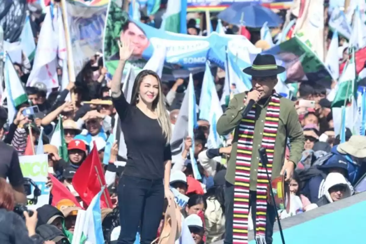 Una mujer saluda con la mano levantada mientras un hombre con sombrero y bufanda colorida habla por micrófono frente a una multitud con banderas y pancartas Una mujer saluda con la mano levantada mientras un hombre con sombrero y bufanda colorida habla por micrófono frente a una multitud con banderas y pancartas