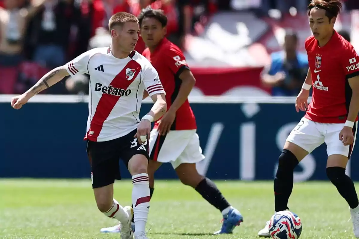 Un jugador de River Plate controla el balón mientras dos rivales con uniforme rojo lo observan durante un partido de fútbol en un estadio. Un jugador de River Plate controla el balón mientras dos rivales con uniforme rojo lo observan durante un partido de fútbol en un estadio.