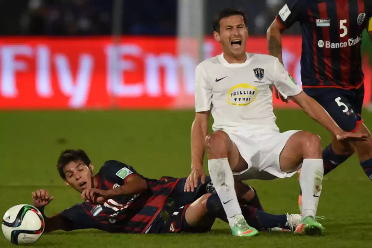 Un jugador con uniforme blanco grita de dolor mientras otro jugador con uniforme azul y rojo está en el suelo tras una jugada durante un partido de fútbol. Un jugador con uniforme blanco grita de dolor mientras otro jugador con uniforme azul y rojo está en el suelo tras una jugada durante un partido de fútbol.